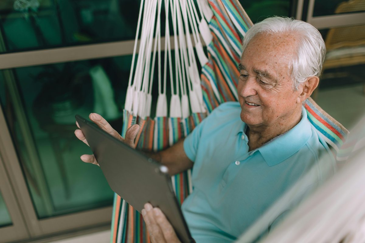 An older man smiling as he uses a tablet on his patio